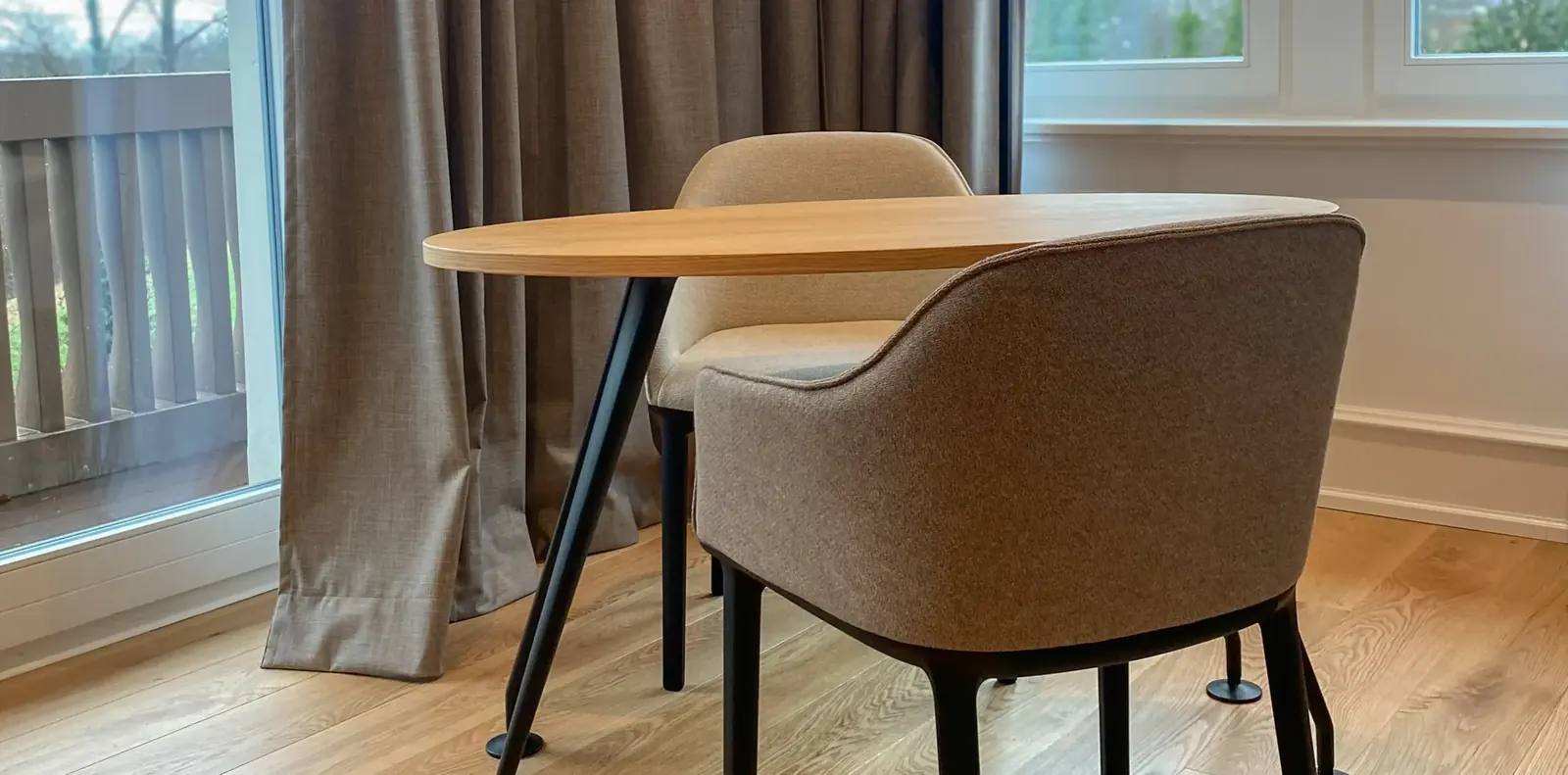 Zwei Polsterstühle in Beige und Braun an einem ovalen Eichentisch vor Balkontür und grauen Vorhängen.