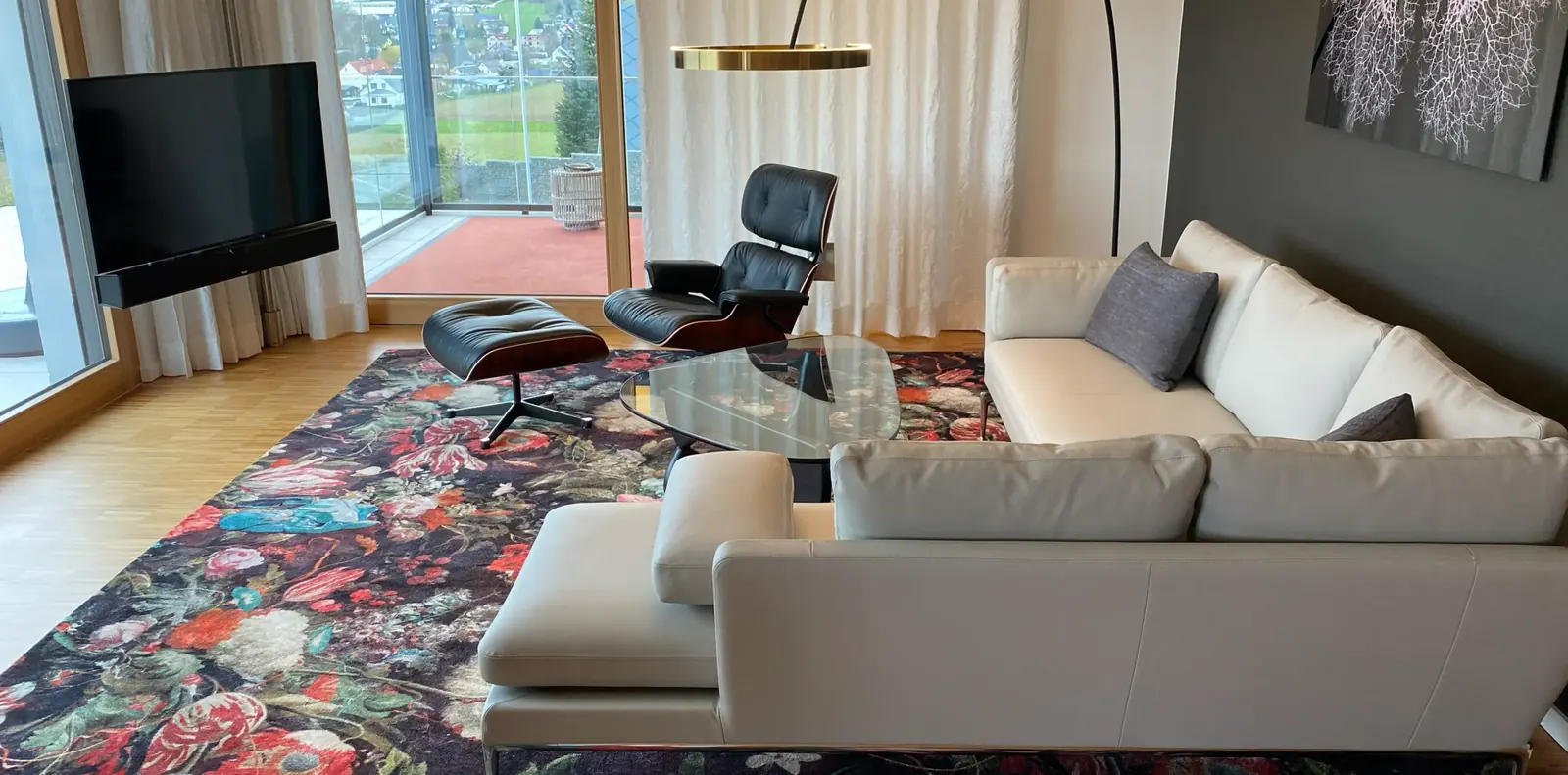 Wohnzimmer mit hellgrauem Ecksofa aus Leder, schwarzem Lounge Chair mit Ottomane und Glastisch auf einem Blumenteppich.