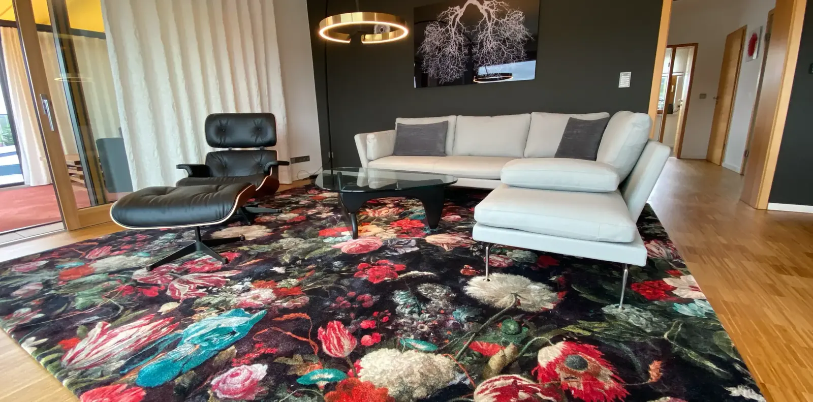 Weißes Ecksofa und schwarzer Lounge Chair mit Hocker um einen Glascouchtisch auf dunklem Teppich mit großem Blumenmuster.