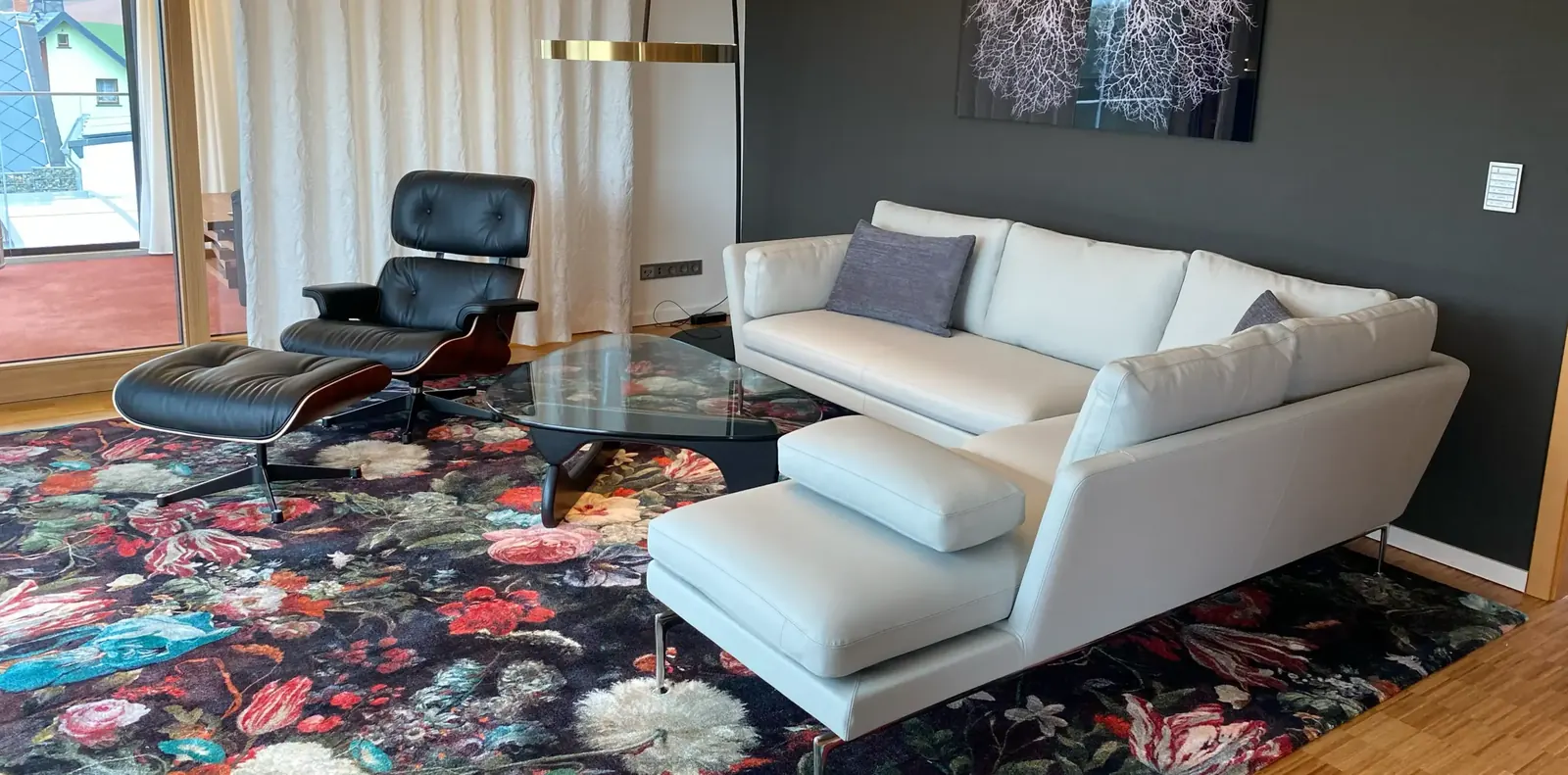 Helles Ecksofa mit grauen Kissen vor anthrazitfarbener Wand, daneben Lounge Chair, Glastisch und Blumenteppich.